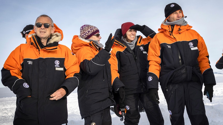 Two men and two women in heavy orange and black winter clothing stand talking on an icy plain.