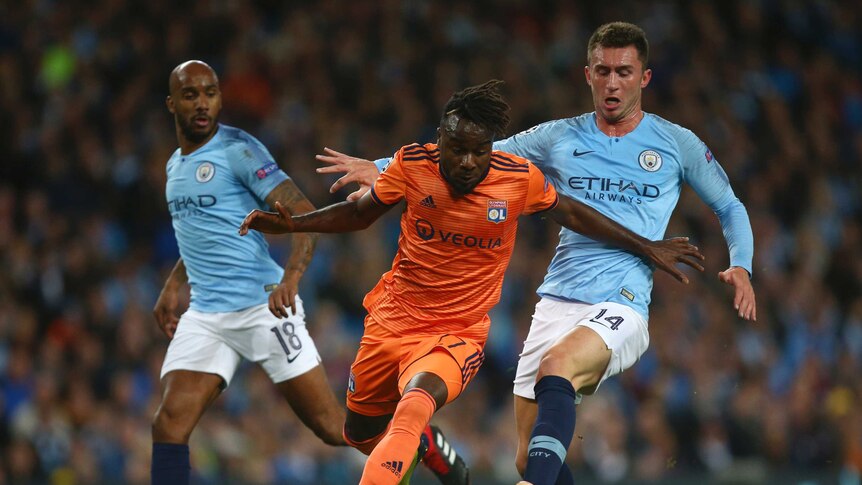 A football player wearing orange stumbles while chasing the ball with two players wearing light blue chasing