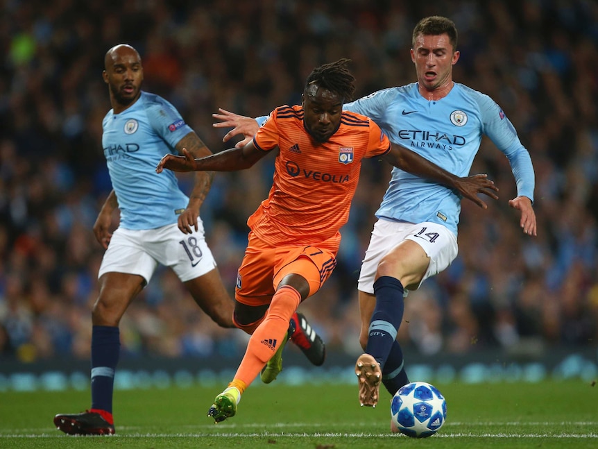 A football player wearing orange stumbles while chasing the ball with two players wearing light blue chasing