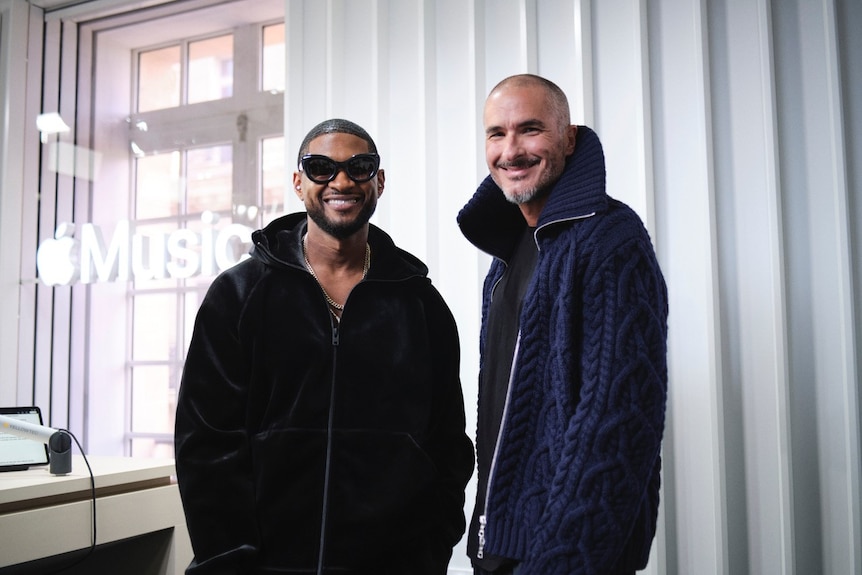 Usher wearing sunglasses and a black top with Zane Lowe in blue, both smiling and standing, hands in pockets