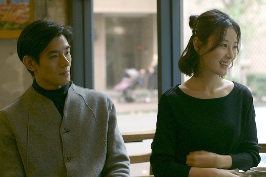 Japanese man in grey coat sits inside cafe next to Japanese woman with dark bob wearing black shirt and smiling.