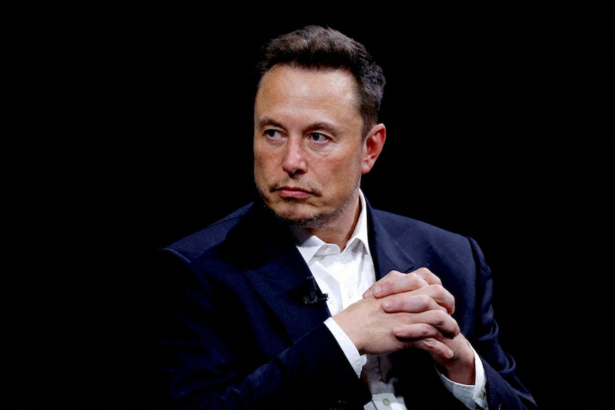 Elon Musk has a serious expression as he looks to his right and clasps his hands together.