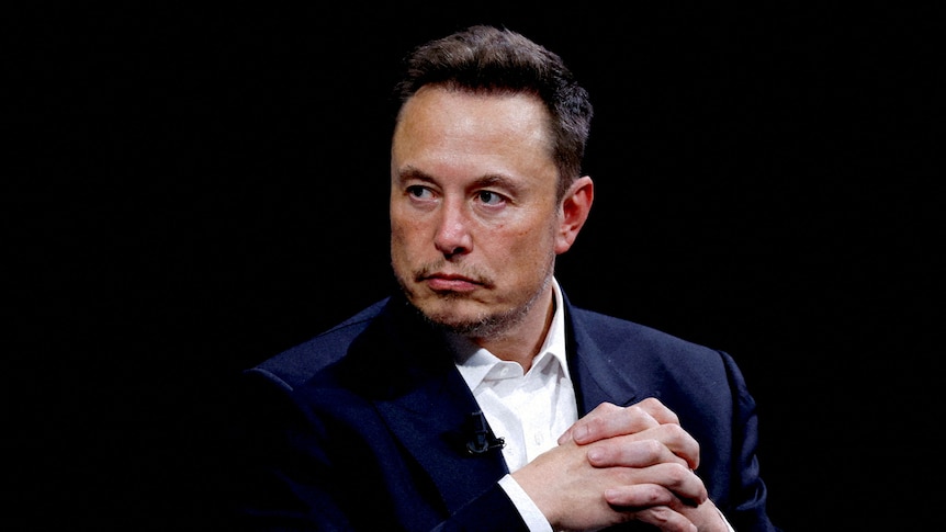 Elon Musk has a serious expression as he looks to his right and clasps his hands together.
