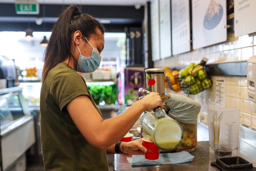 A worker wearing a mask pours a drink into a cup.