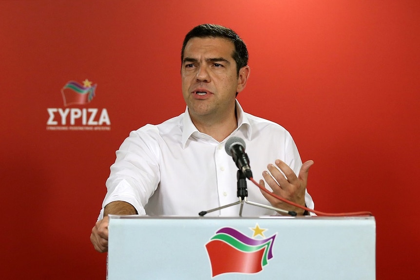 A man speaks into a microphone mounted on a lectern in front of the red political banner of a Greek political party.