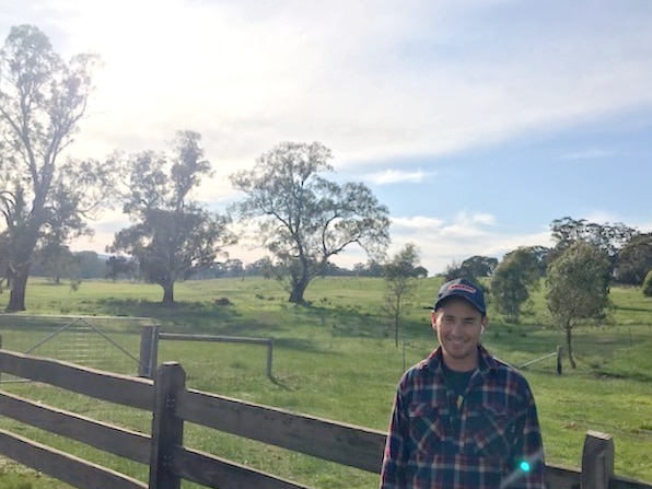A young man with flannelette shirt at a fence on a green, lush farm. He is smiling and the sun is in the background.