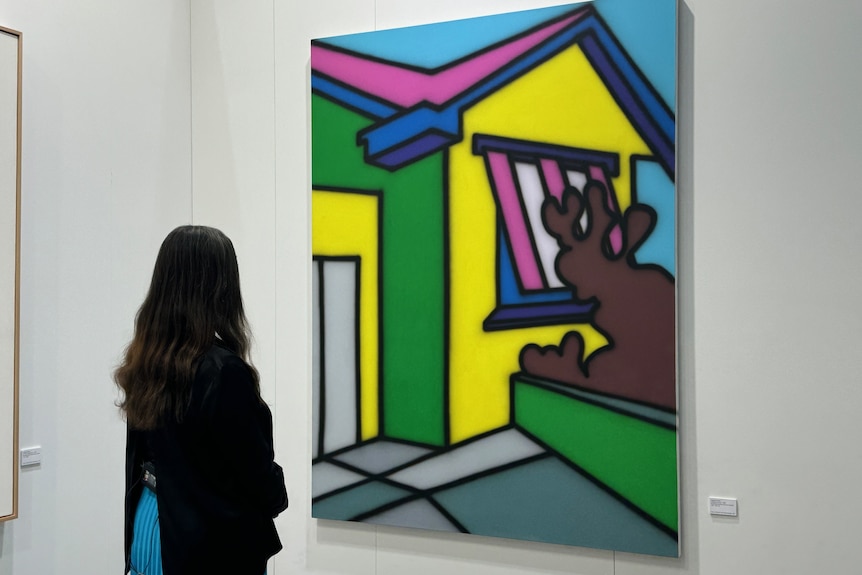 Una mujer con cabello largo y oscuro mira una pintura gráfica brillante de una casa en una galería de arte.