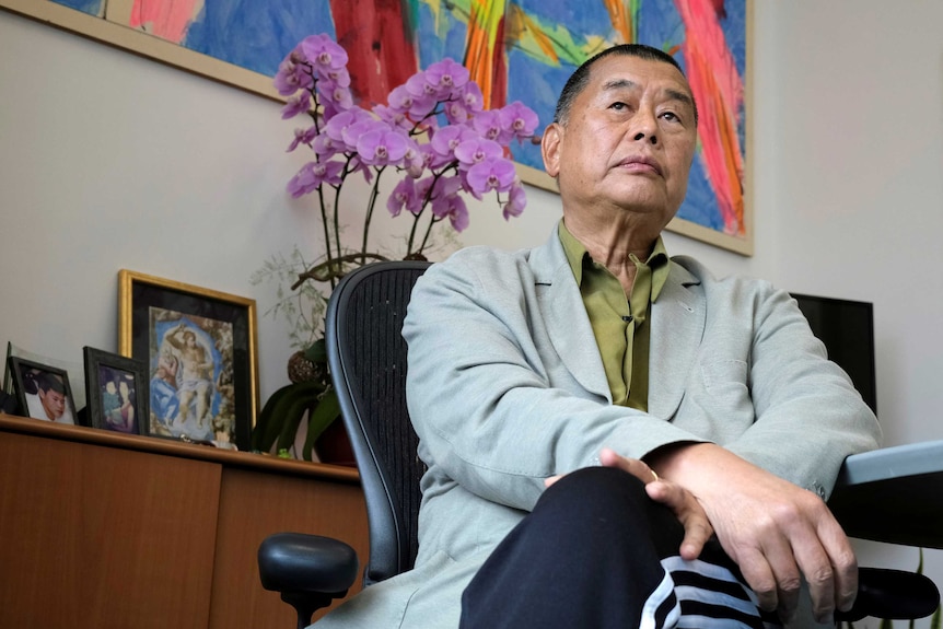 A man in a green shirt and grey blazer sits looking contemplative in front of a colourful painting and potted orchid