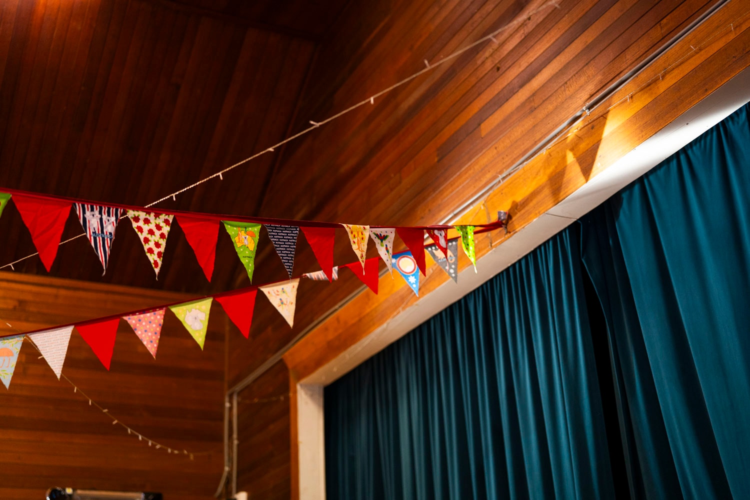 Small red and yellow flags float in a community hall with wooden panels and a teal velvet curtain