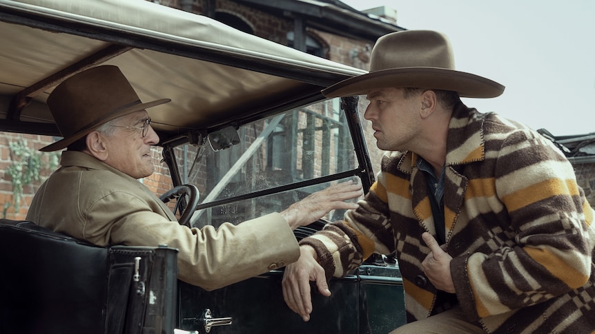 A film still showing an older man wearing a cowboy hat sitting in a vehicle talking to a younger man in a hat and striped coat