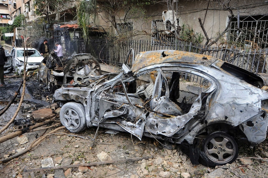 A burnt out car sits in the middle of road covered in debris