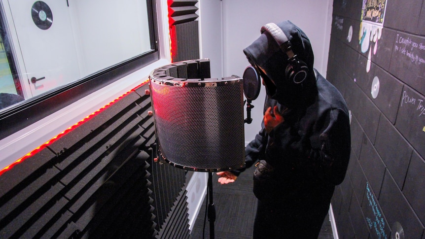 Renski stands in front of a microphone in the recording booth wearing a dark hoodie.
