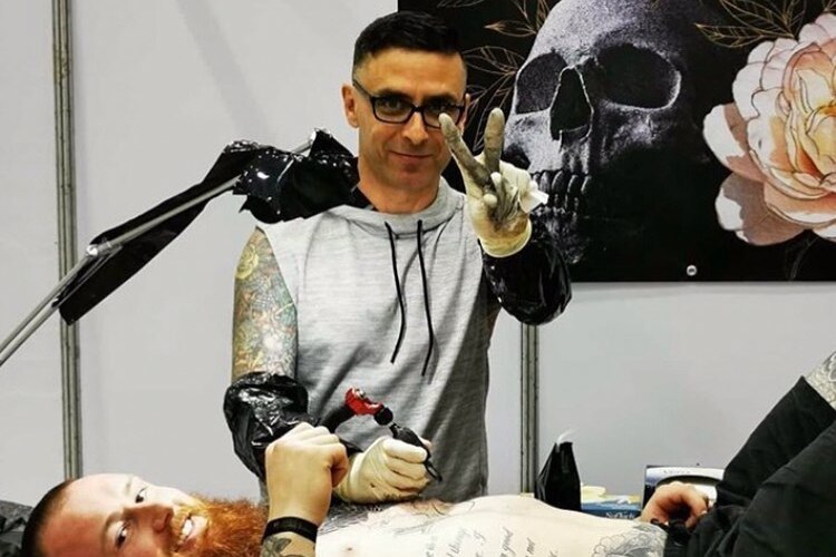 Chris Rand gives a peace sign while tattooing a man lying on his back. 