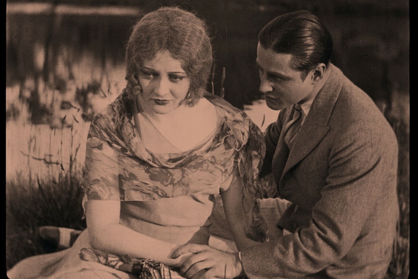 A grainy still image from a silent film showing a man and a woman sitting beside a lake looking solemn.