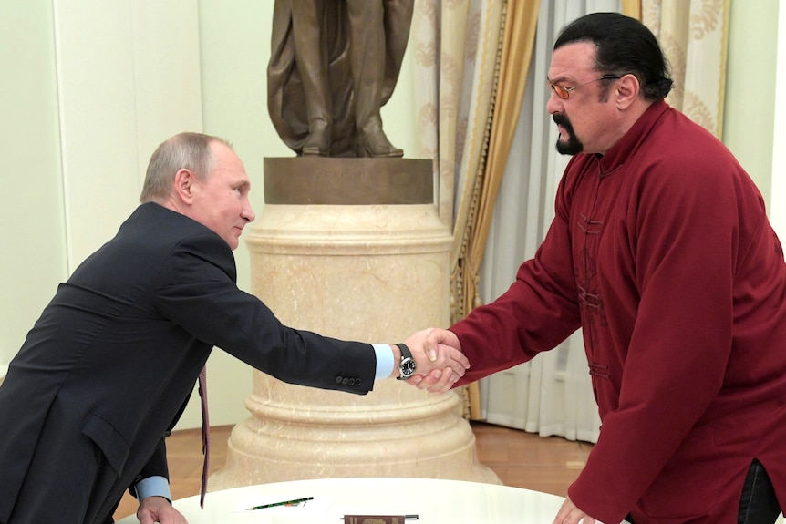 A short man in a suit shakes the hand of a taller man wearing red in front of a table in a room