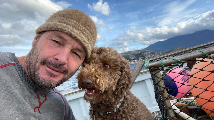 A man with a stubble beard and beanie poses with a poodle on a fishing boat.
