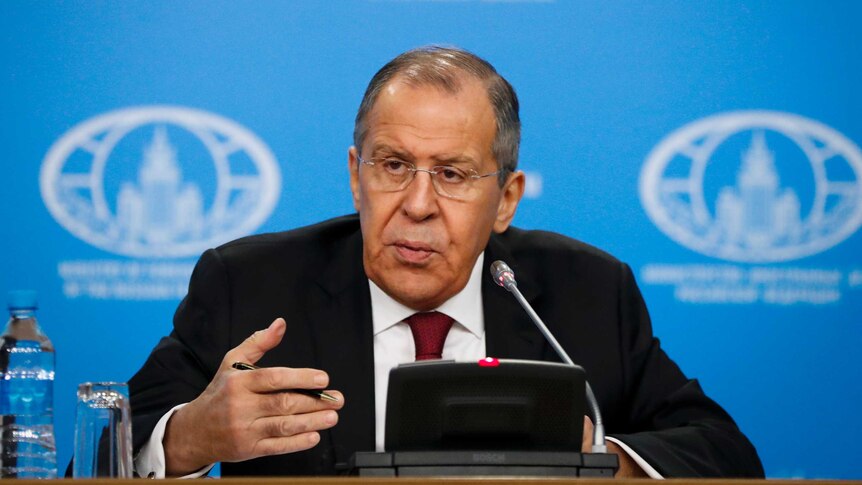 Sergey Lavrov sits behind a lectern with a microphone, gesturing with one hand while holing a pen. There is a blue screen behind