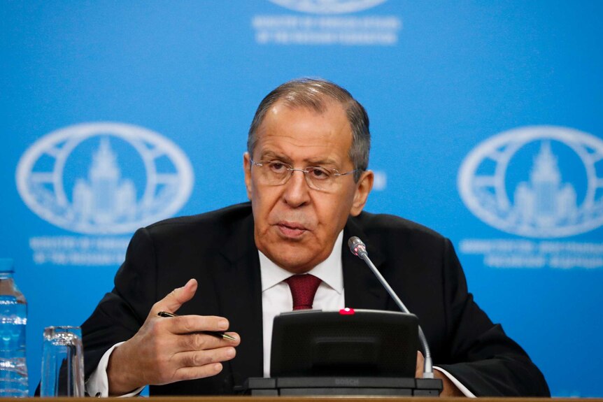 Sergey Lavrov sits behind a lectern with a microphone, gesturing with one hand while holing a pen. There is a blue screen behind