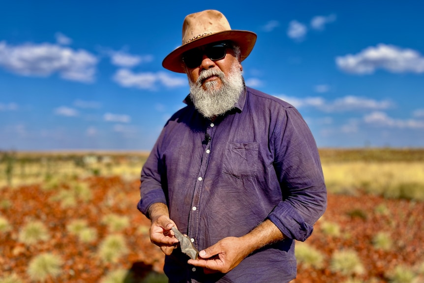 A man wearing a broad-brimmed hat stands in the outback on a sunny day.