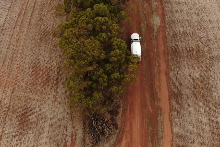 An aerial shot of a water truck on a brown dirt road, with rural land on either side. 