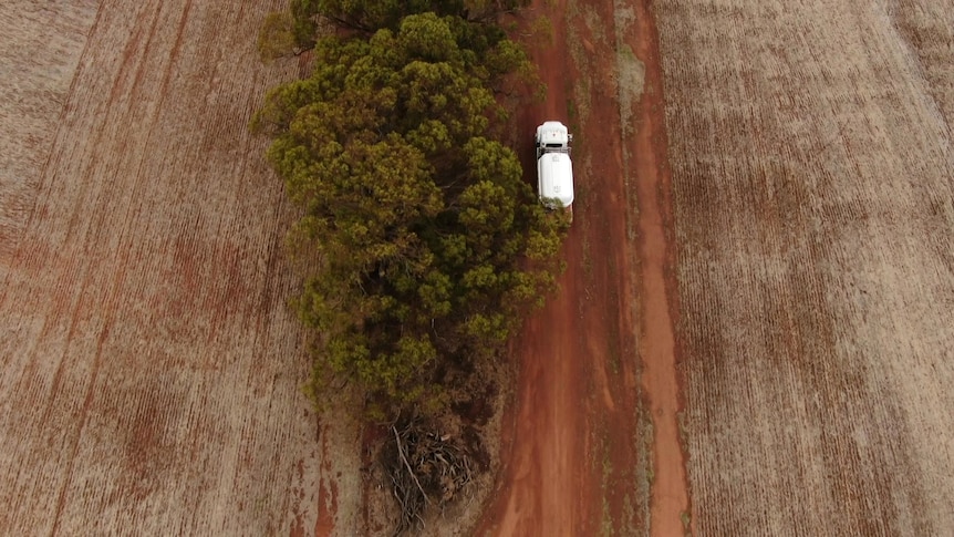 An aerial shot of a water truck on a brown dirt road, with rural land on either side. 