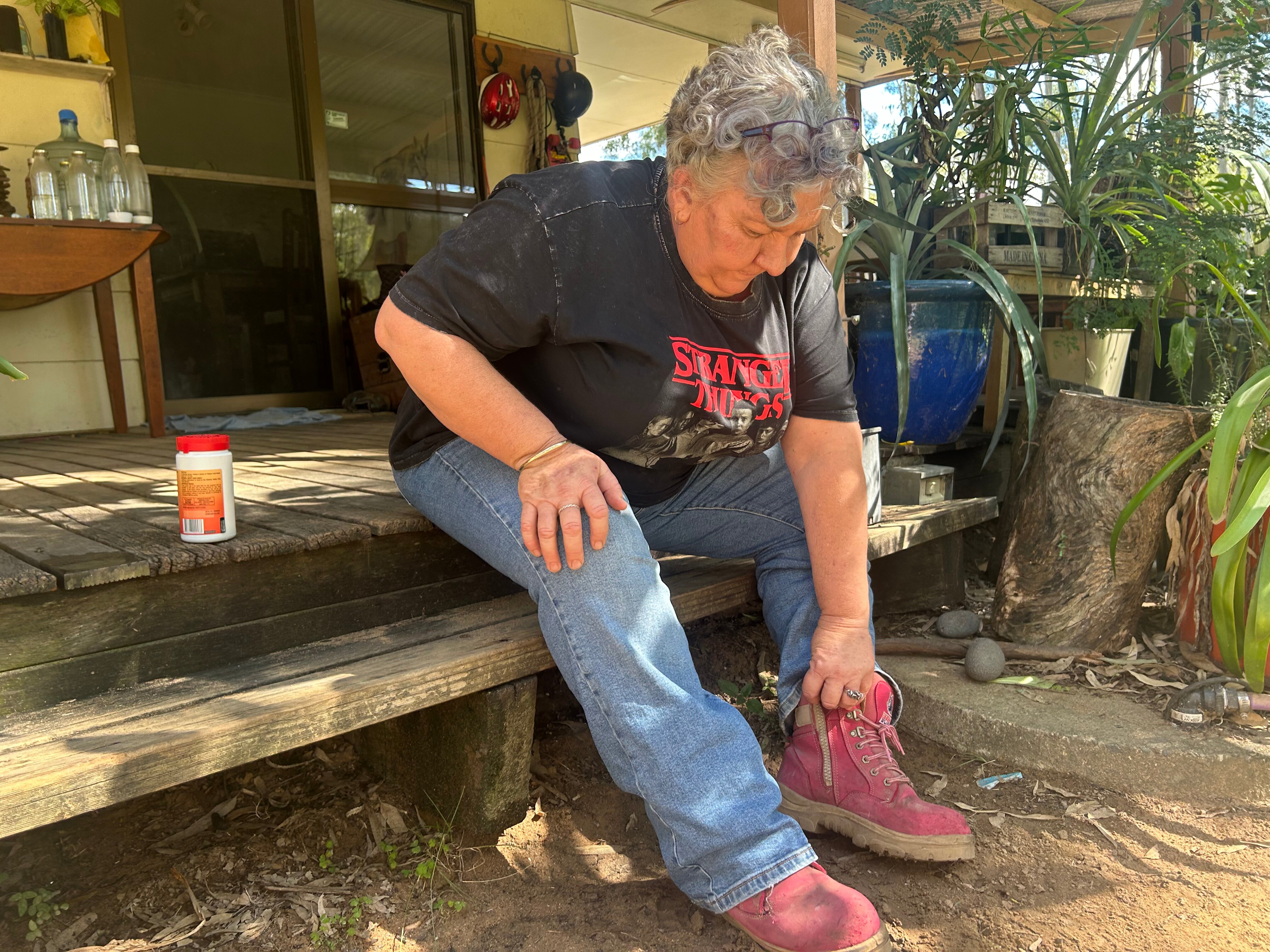 A woman wearing a t-shirt and jeans sits on the front step and puts on pink sneakers.