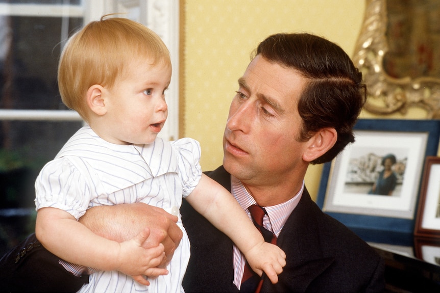 A young Prince Harry being held by his father Prince Charles who is looking at him with a serious expression