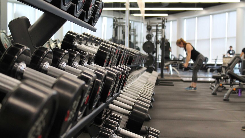 A rack of weights in a gym is in focus while a woman lifts dumbbells in the background