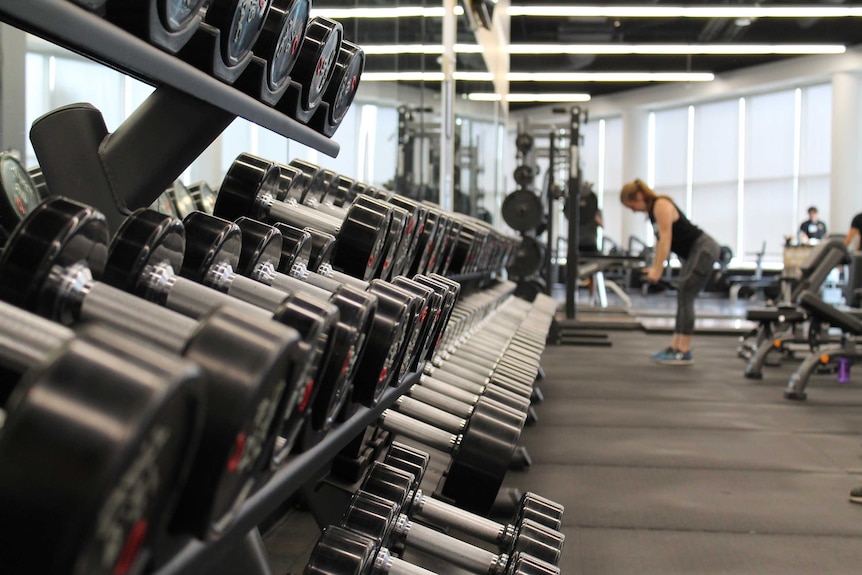 A rack of weights in a gym is in focus while a woman lifts dumbbells in the background