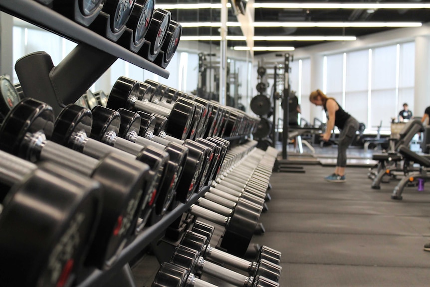 A rack of weights in a gym is in focus while a woman lifts dumbbells in the background.