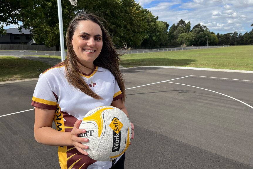 A young woman with brown hair, wearing a white, yellow and brown hawks netball top, holding a netball. She's standing on a court