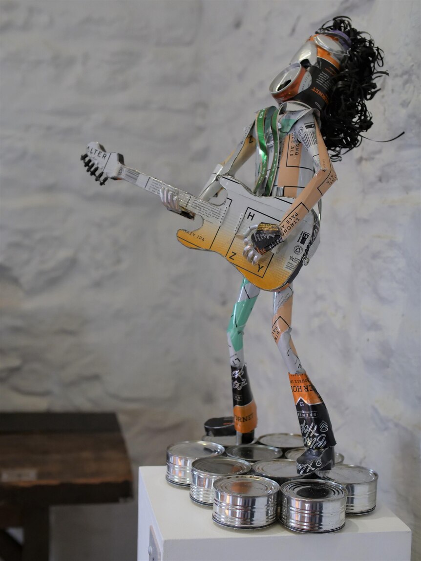 A rock star sculpture made from aluminium cans, standing on a collection of shortened tin cans.