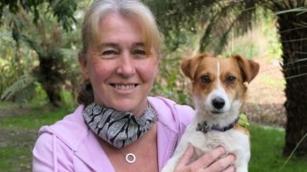 A woman with blonde hair smiles at the camera while holding a small white and brown dog, they are in a garden.