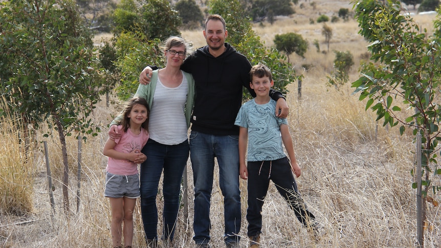 A young family stands with their arms around each other as they smile into the camera, standing in front of trees