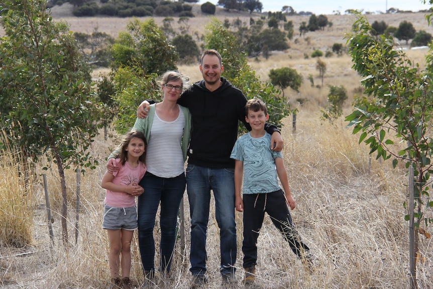 A young family stands with their arms around each other as they smile into the camera, standing in front of trees
