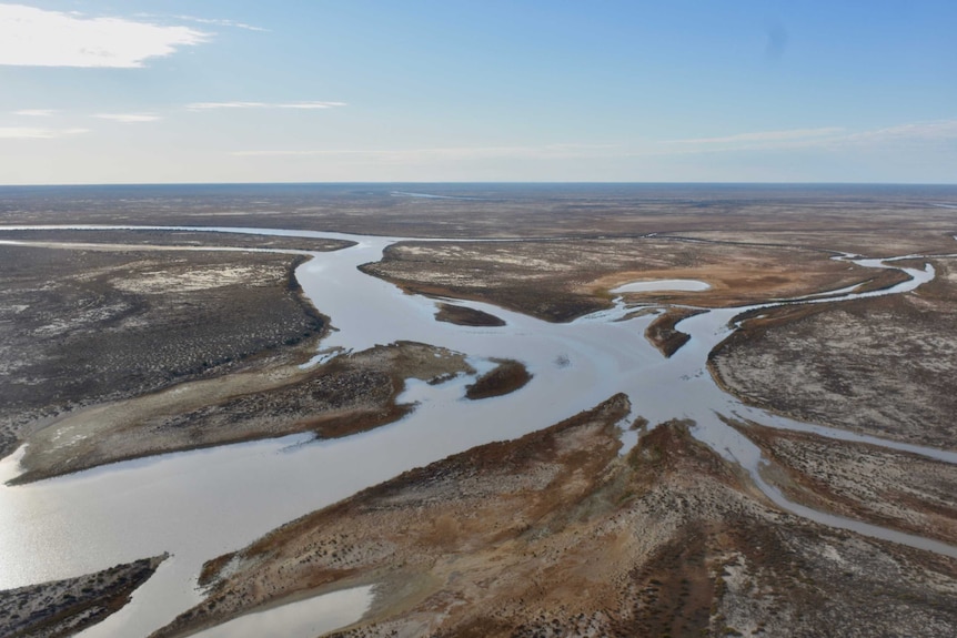 A blue-green coloured creek winds through a vast, open and arid countryside with the horizon stretching far into the distance