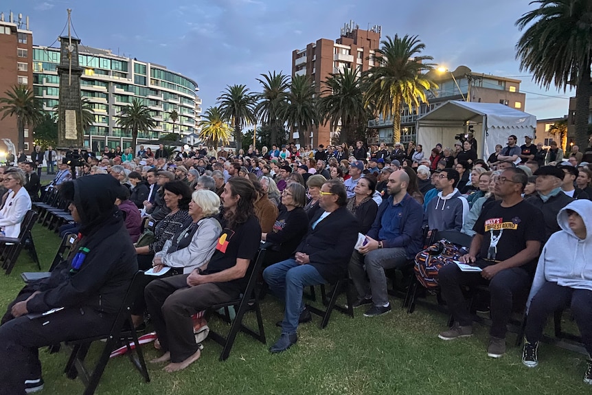A large gathering of people sit on fold out chairs on the grass on the St Kilda foreshore in the dawn light.