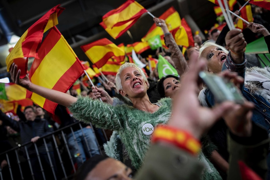 A woman in a furry green jumper and short blonde hair waves a yellow and red striped Vox flag during a rally.