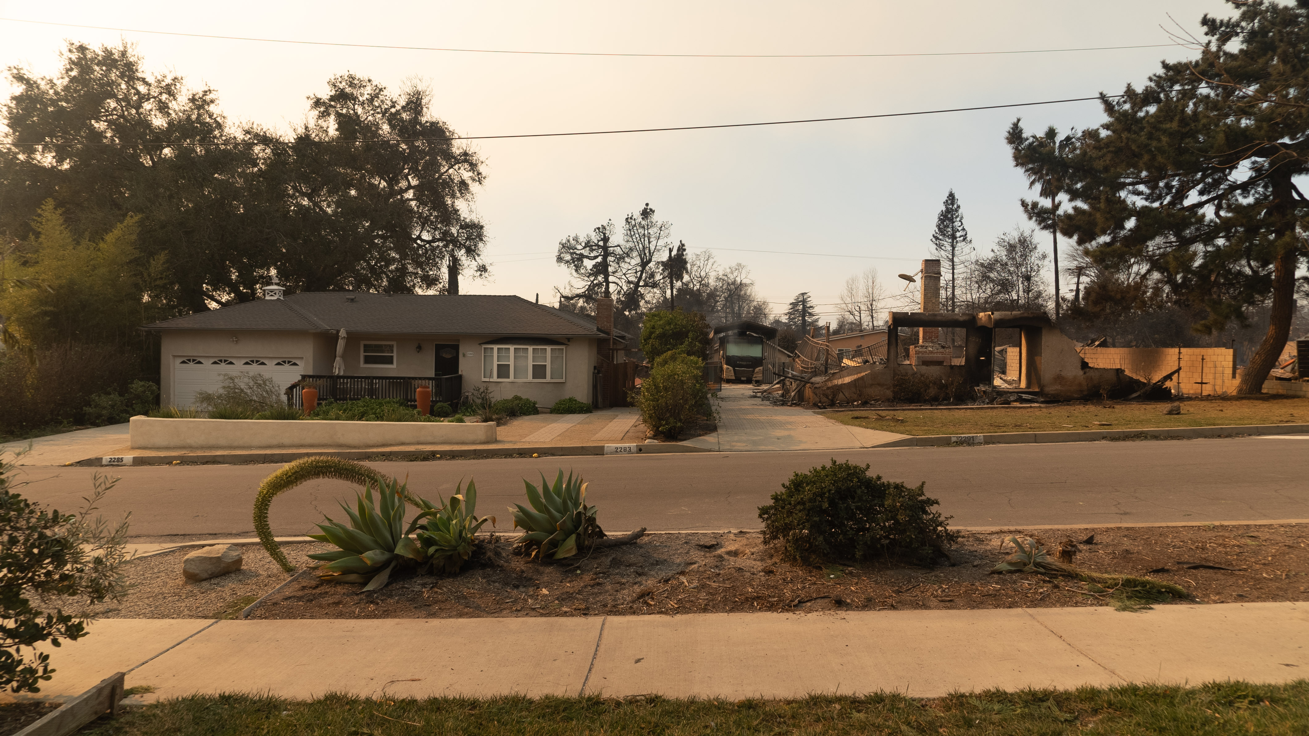 A view of a street, where one house is completely burnt down as smoke clouds the air