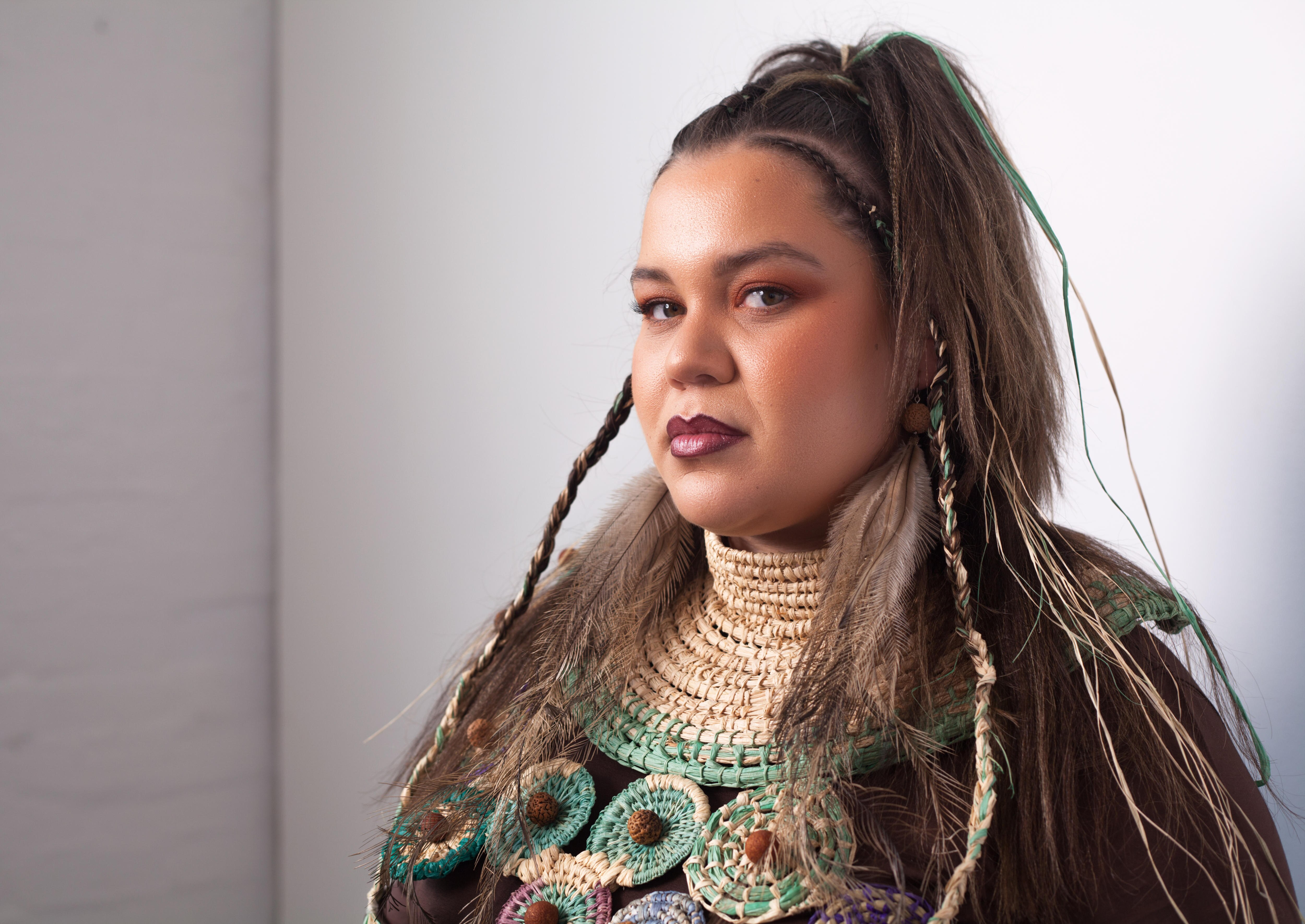 An Indigenous woman in a plain room, looking at the camera with a serious expression
