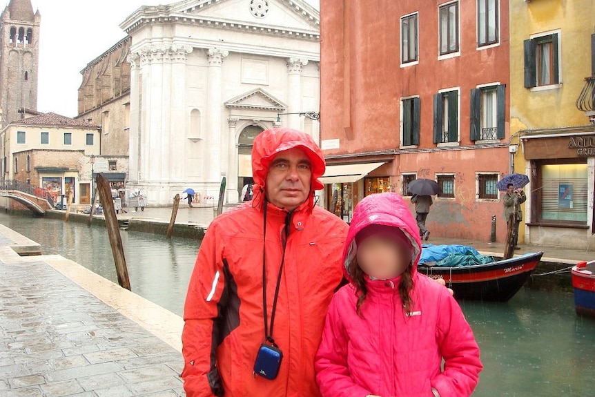 Oleg Kouzmine in front of a canal and ornate buildings with a girl, both wearing rain jackets.
