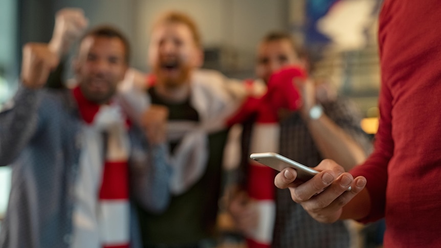 A man distracted by his phone while his friends enjoy a sporting match behind him.