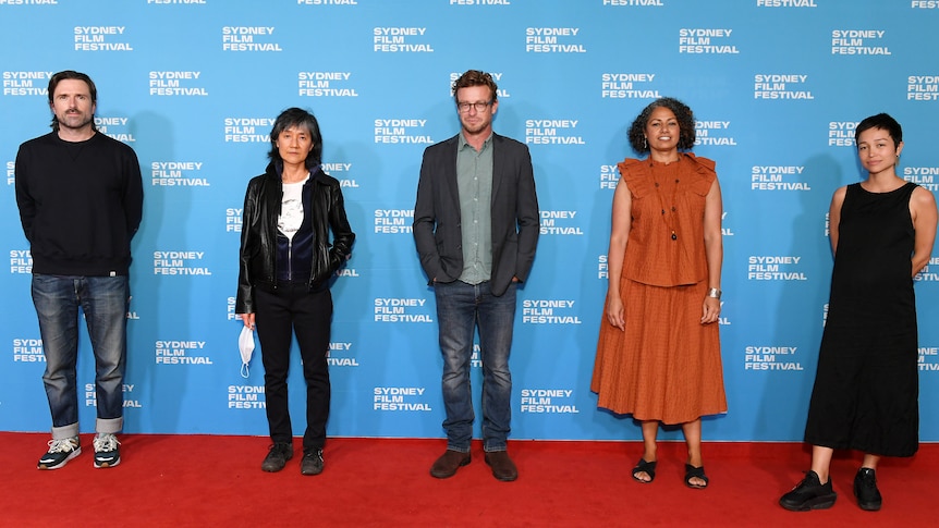 Five people stand in a line on red carpet with blue logo-covered wall behind them. 