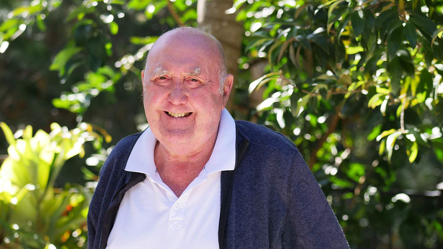 elderly man with with white shirt and blue zip up jumper smiles at the camera.