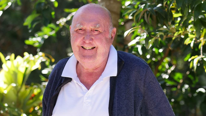 elderly man with with white shirt and blue zip up jumper smiles at the camera.