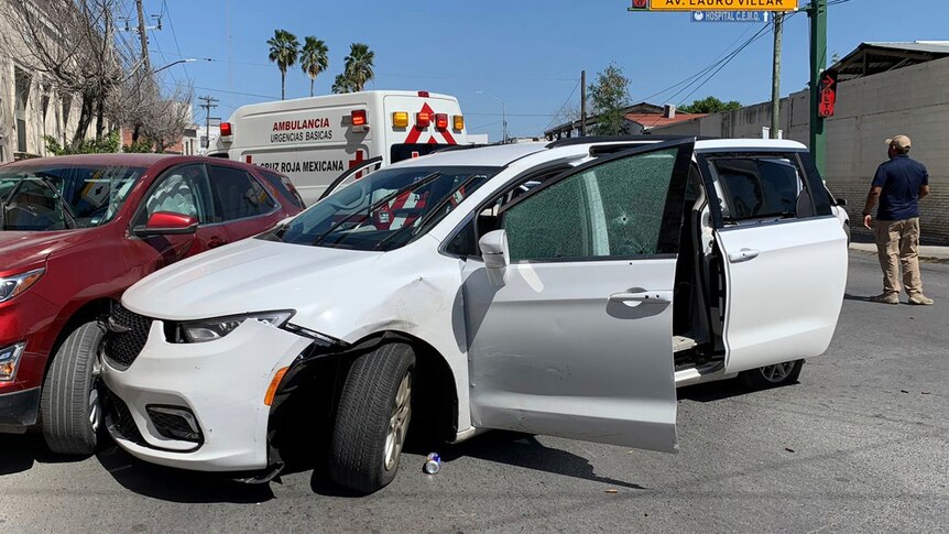 A white mini-van shows crash damage and bullet damage including crazed window glass.