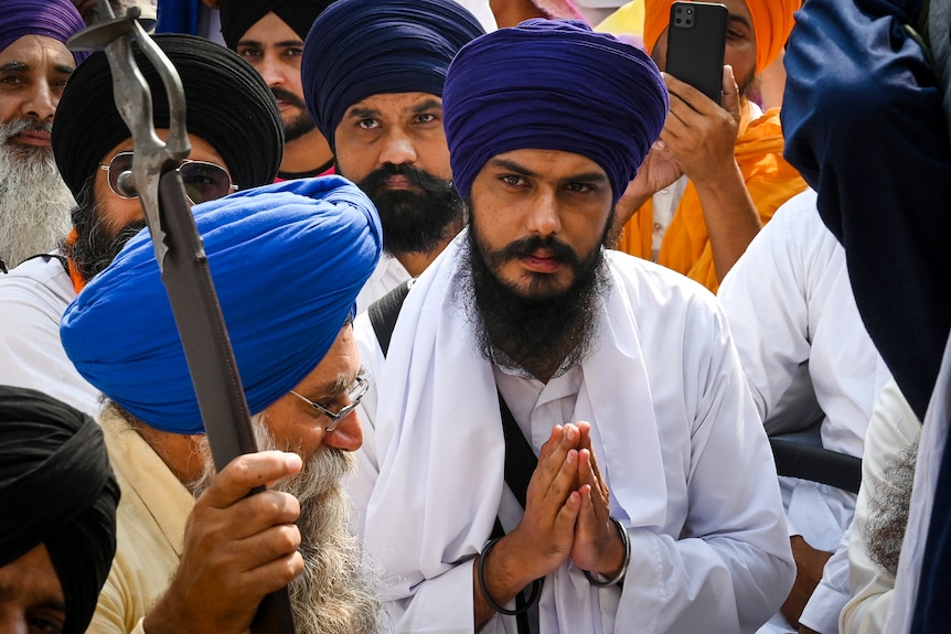 A preacher wearing a white robe and blue turban holds his hands in a prayer position