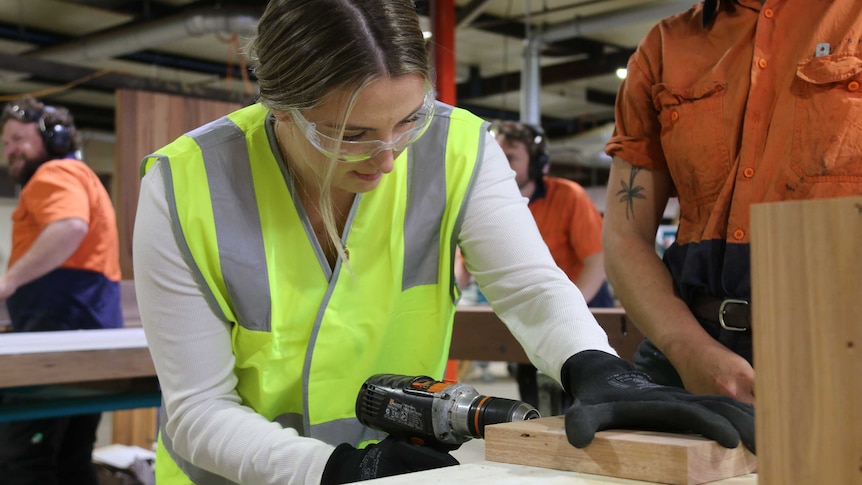 A young woman in a workshop holds a piece of wood to a table as she begins to drill it.