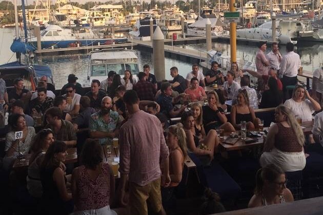 A crowd of people are packed into tables drinking and eating alongside a marina.