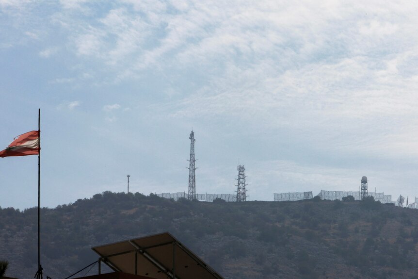 Metal towers and wire fencing sit atop a sparse-looking hillside under a dull blue sky.